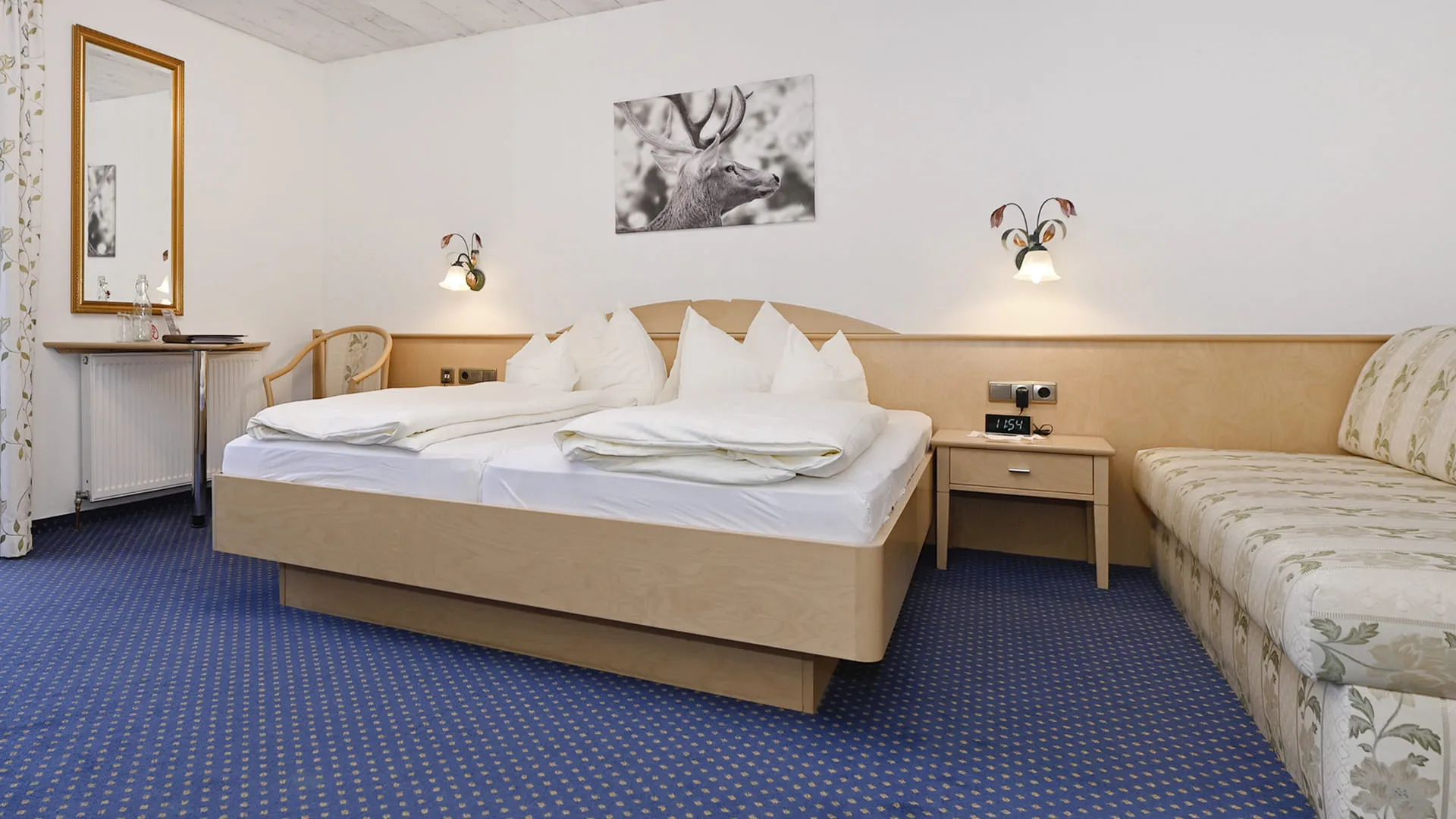 Hotelzimmer mit Doppelbett, Nachttisch und Sofa, blauser Teppich und Spiegel