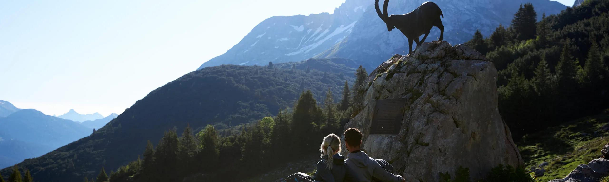 Zwei Kinder sitzen auf einem Felsen mit Ziegenbock im Lechweg bei Alpengipfeln.