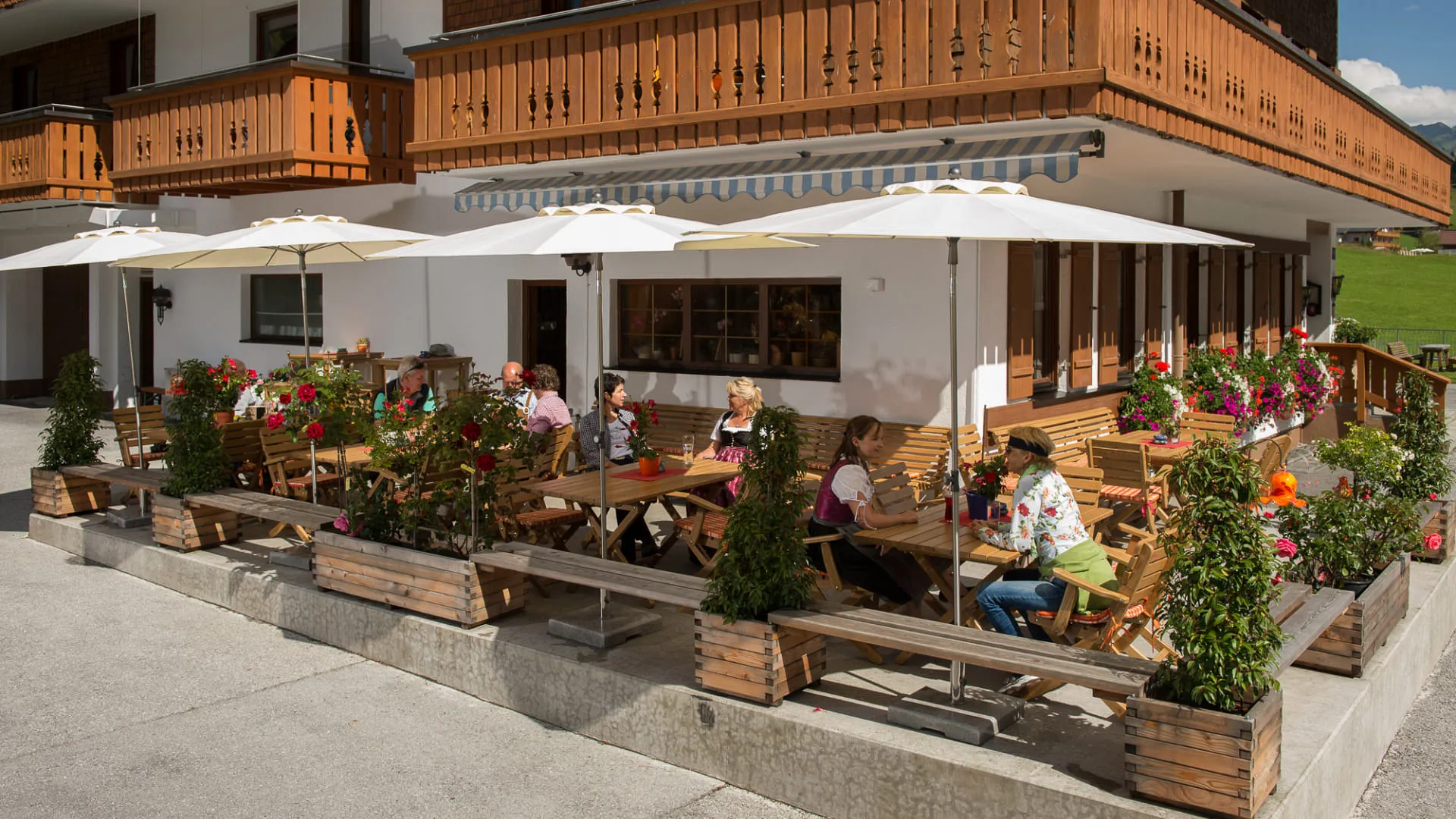 Terrasse eines Hauses mit Gästen unter Sonnenschirmen, Blumen und Tischen.